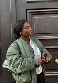 Giacca bomber verde con polsini a coste, indossata sopra una camicia bianca. Tiene degli occhiali da sole e un telefono, su uno sfondo di legno scuro.