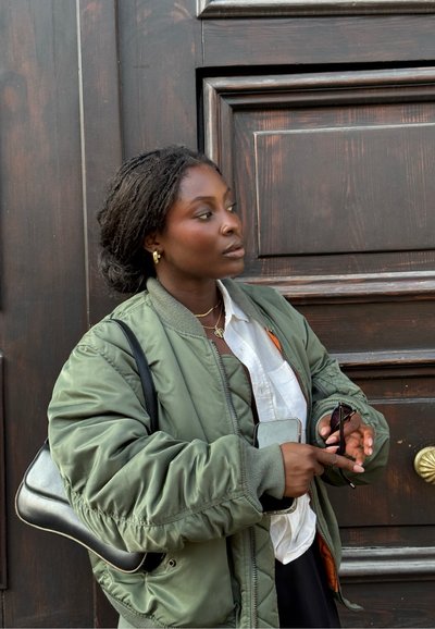 Cazadora bomber verde con puños acanalados, sobre una camisa blanca. Sosteniendo unas gafas de sol y un teléfono, contra un fondo de madera oscura.