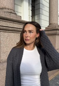 Grå strikket cardigan med ribbestrikket krage, båret over en hvit tettsittende skjorte. Cardiganen har teksturert stoff og lange ermer.