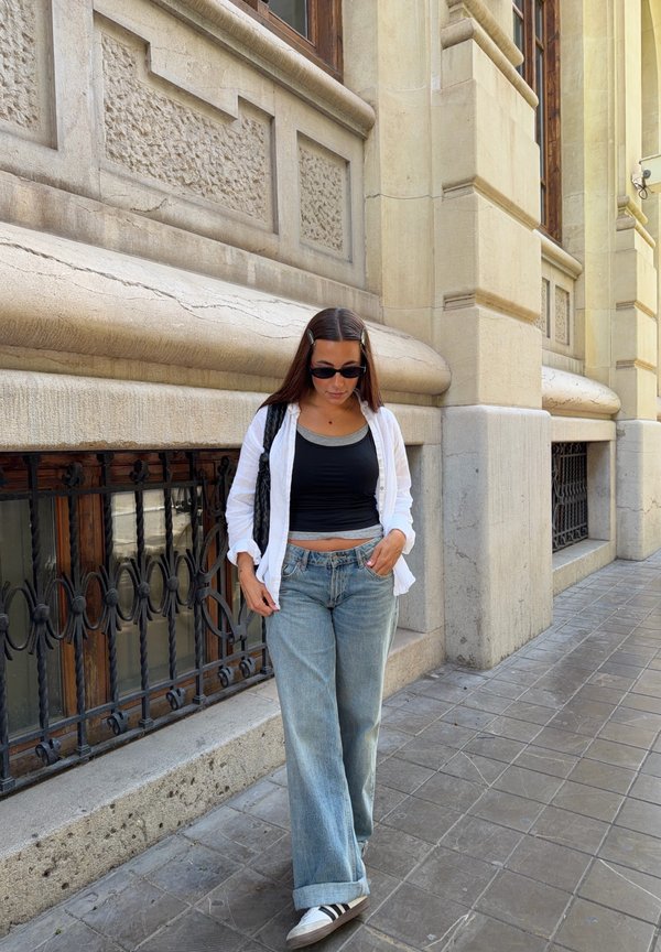 Žena nosící sluneční brýle, černý crop top, bílou košili a džíny kráčí po městském chodníku vedle ozdobné kamenné budovy s kovovými mřížemi oken.
