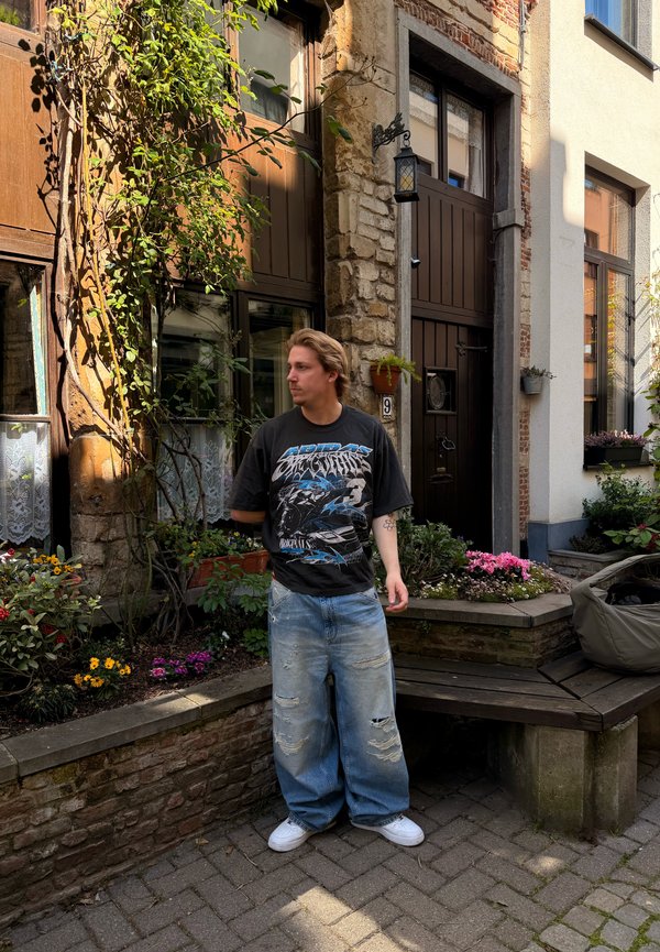Un jeune homme se tient près d'un bâtiment en pierre avec des portes et fenêtres en bois, entouré de fleurs et de verdure dans une petite cour.