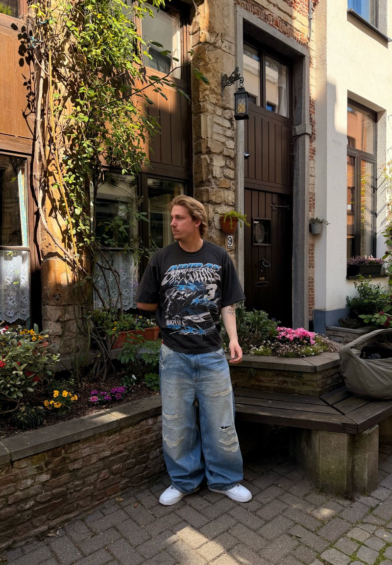 Un jeune homme se tient près d'un bâtiment en pierre avec des portes et fenêtres en bois, entouré de fleurs et de verdure dans une petite cour.