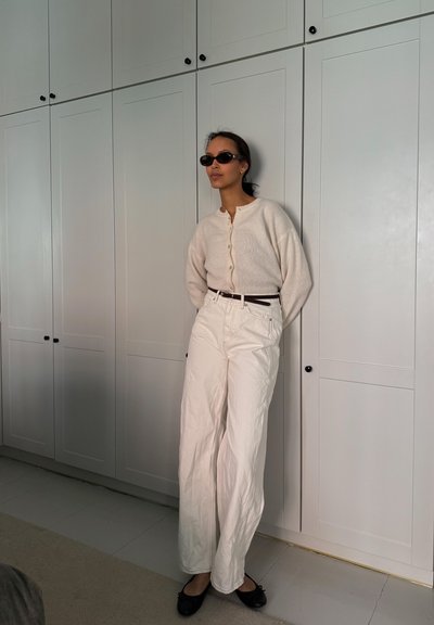 Cárdigan crema de botones, pantalones de pierna ancha blancos de cintura alta, cinturón negro y bailarinas negras. La modelo lleva gafas de sol oscuras con un diseño minimalista.