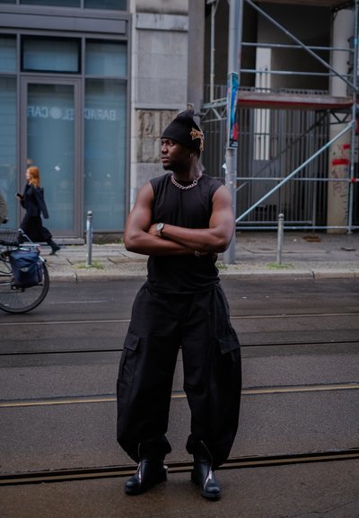 Pantalones cargo negros, camiseta negra sin mangas, gorro negro con adornos, collar de cadena plateada, de pie con los brazos cruzados en una calle.