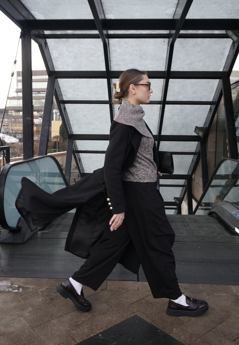 Femme en manteau noir et pantalon large qui marche d'un pas rapide devant des escalators dans un cadre urbain moderne avec un toit en verre, tenant une pochette noire.