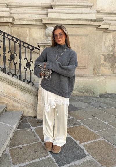 Suéter gris de punto de gran tamaño con un escote alto, superpuesto a un conjunto blanco de satén con encaje, completado con zapatos tipo slip-on beige.