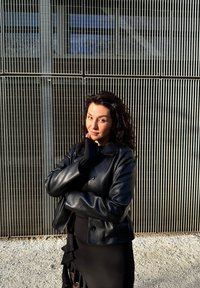 Giubbotto corto in pelle nera con grandi bottoni, abbinato a un vestito nero con rouches. I lunghi e ricci capelli scuri sono acconciati in modo naturale.