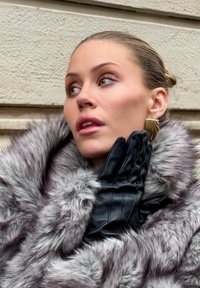 Szary futrzany płaszcz o miękkiej fakturze i długich włóknach, noszony na czarnych skórzanych rękawiczkach z pionowymi szwami i błyszczącym wykończeniem.