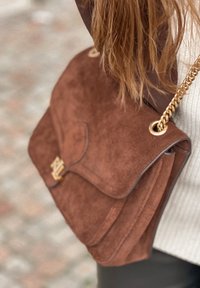 Sac à main en daim marron avec une fermeture à rabat et une bandoulière chaîne dorée. Le sac présente des accents texturés et un détail de logo à l'avant.