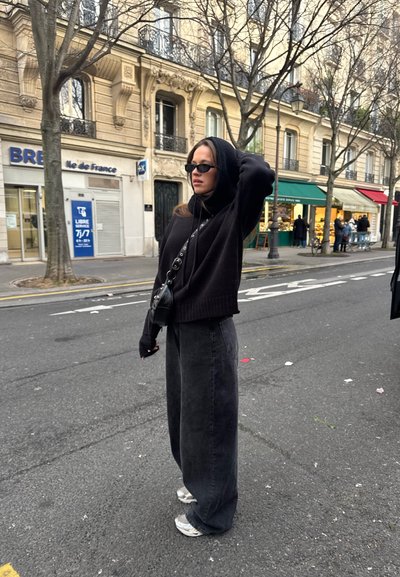 Sudadera negra con capucha, jeans anchos negros, zapatillas plateadas y un pequeño bolso cruzado negro. Las gafas de sol completan el conjunto. Fondo de escena urbana.