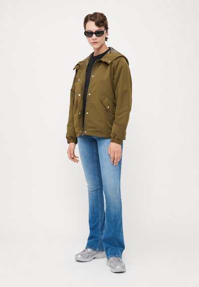 Chaqueta verde oliva con capucha y cierres a presión, con dos bolsillos frontales, combinada con jeans de campana azul claro y zapatillas deportivas grises.