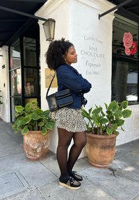 Schwarze Handtasche mit glänzender Oberfläche, Jeansjacke, Leopardenmuster-Rock, schwarze Strumpfhosen und Schuhe mit Tiermuster, vor einer weißen Wand.