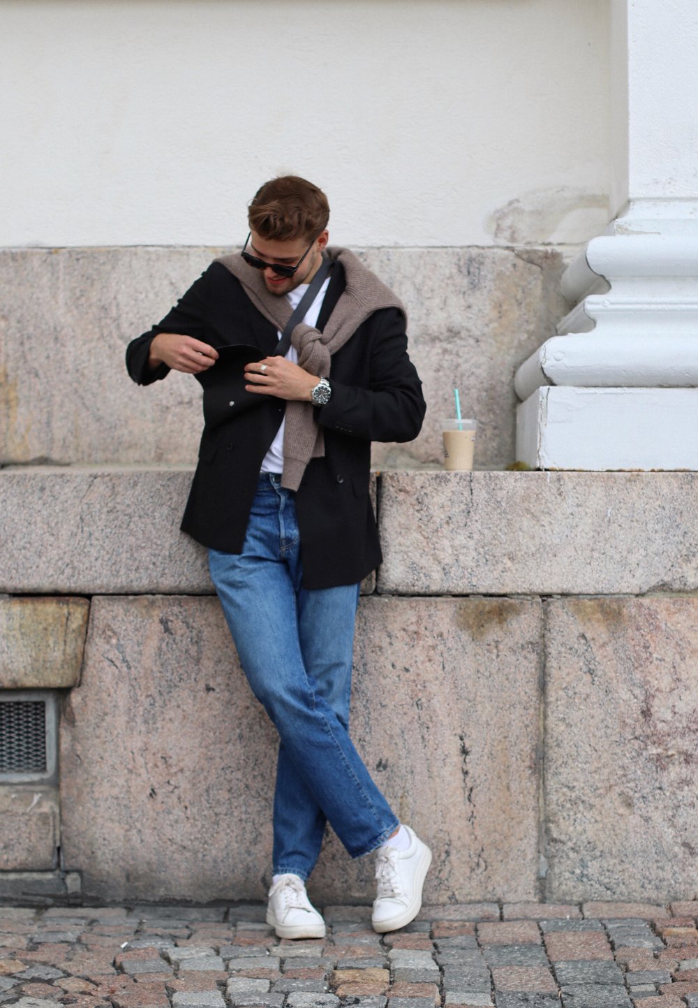 Sort blazer over hvid skjorte, beige strikketrøje draperet, blå jeans, hvide sneakers. Holder en kopi tæt på en stenmur.