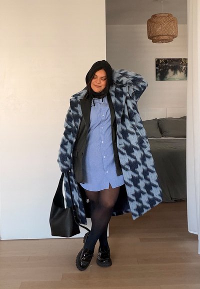 Mujer con un abrigo azul estampado sobre un vestido azul, medias negras y zapatos, llevando un bolso negro, de pie en la puerta de un dormitorio.