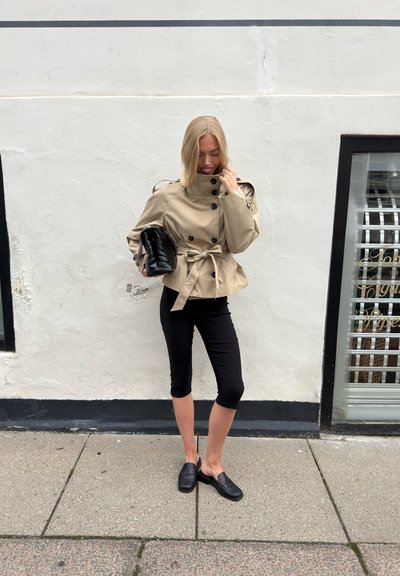 Mujer con chaqueta beige ceñida con cinturón, leggins negros cortos y zapatos negros sin cordones, sosteniendo un bolso acolchado negro, de pie en la acera junto a una pared blanca.