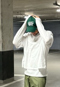 Groene pet met witte tekst, witte lange mouw shirt en olijfgroene broek in een parkeergarage met betonkolommen.