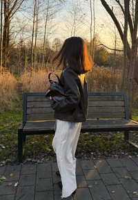 Giacca di pelle nera, pantaloni bianchi a gamba larga con orlo sfrangiato, tiene in mano una piccola borsa nera, in piedi su un sentiero di pietra accanto a una panchina di legno.