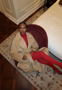 Abrigo de lana color beige con solapas anchas, llevado sobre un suéter de cuello alto rojo, combinado con medias rojas. Sobre una silla mullida de color burdeos, con una alfombra estampada de fondo.