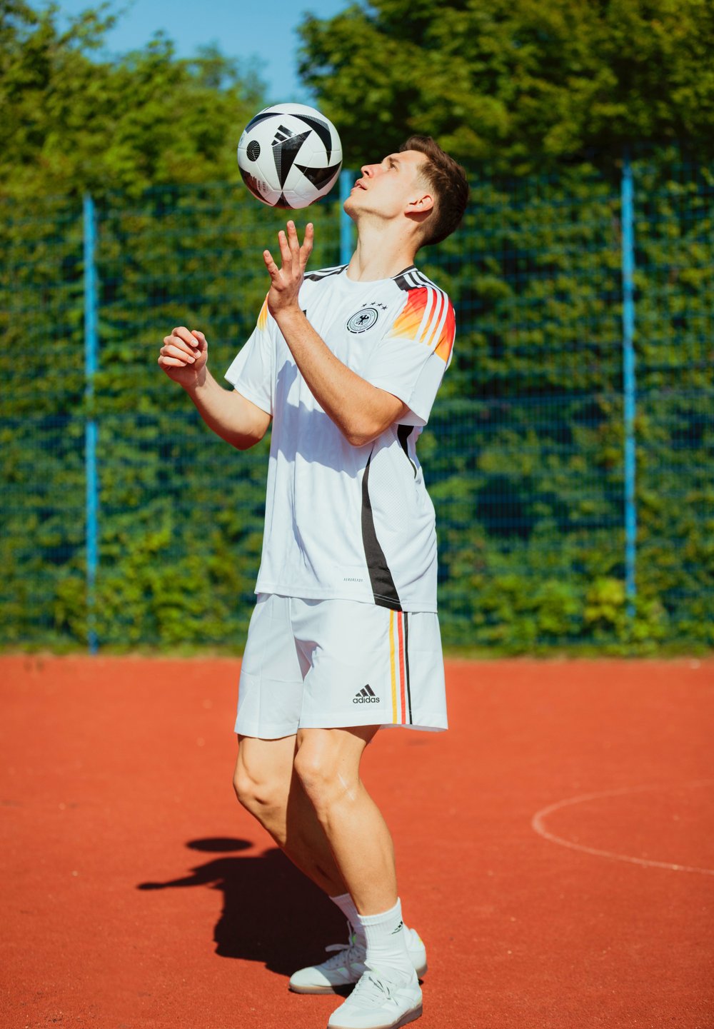 Bela športna uniforma Adidas z oranžnimi, črnimi in rdečimi črtami, črno-bela nogometna žoga na rdečem igrišču, obdanem z zelenjem.