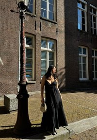Frau in einem schwarzen ärmellosen Kleid, die eine kleine geflochtene Tasche hält, steht auf einer sonnigen Straße in der Nähe eines dekorativen Laternenpfahls und der Fenster eines Ziegelgebäudes.