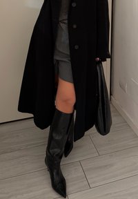 Fekete térdig érő csizma hegyes orral, szürke szoknyával és fekete kabáttal párosítva. A kabáton nagy gombok és testhezálló sziluett található.