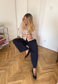 Cardigan tricotat bej cu nasturi, pantaloni largi negri, și balerini negri cu fundițe. Pardoseală din lemn deschis și un fundal minimalist.