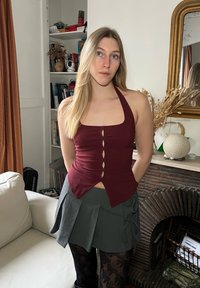 Top halter bordeaux com detalhes em botão, combinado com uma saia plissada cinza com laços laterais. Em fundo de interior de casa.