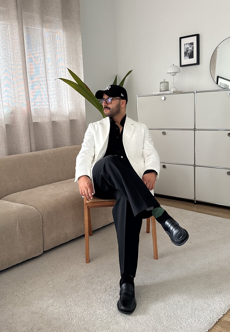 Hvid blazer over en sort skjorte, sorte bukser og sorte loafers. Sidder på en træstol med en plante, lyse gardiner og et moderne skab.