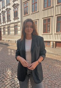 Juodas per didelis švarkas, baltas prigludęs marškinėlis, pilkos džinsus. Akmenimis grįsta gatvė. Šiltas dienos šviesa pabrėžia tekstūras. Minimalūs aksesuarai, laisvas kirpimas.