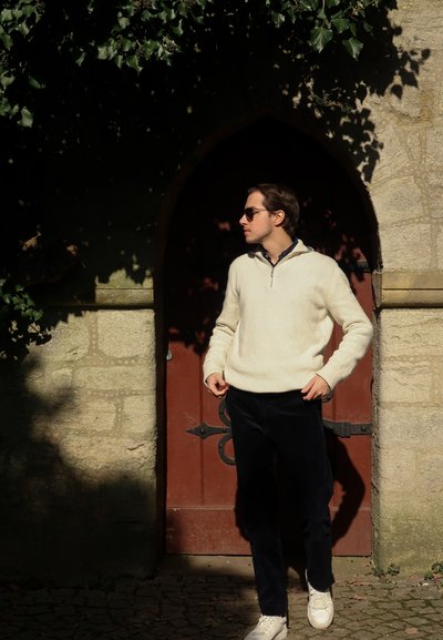 Suéter de punto crema con cuello de media cremallera, combinado con pantalones de pana en color azul marino y zapatillas blancas. Unas gafas de sol oscuras completan el look.