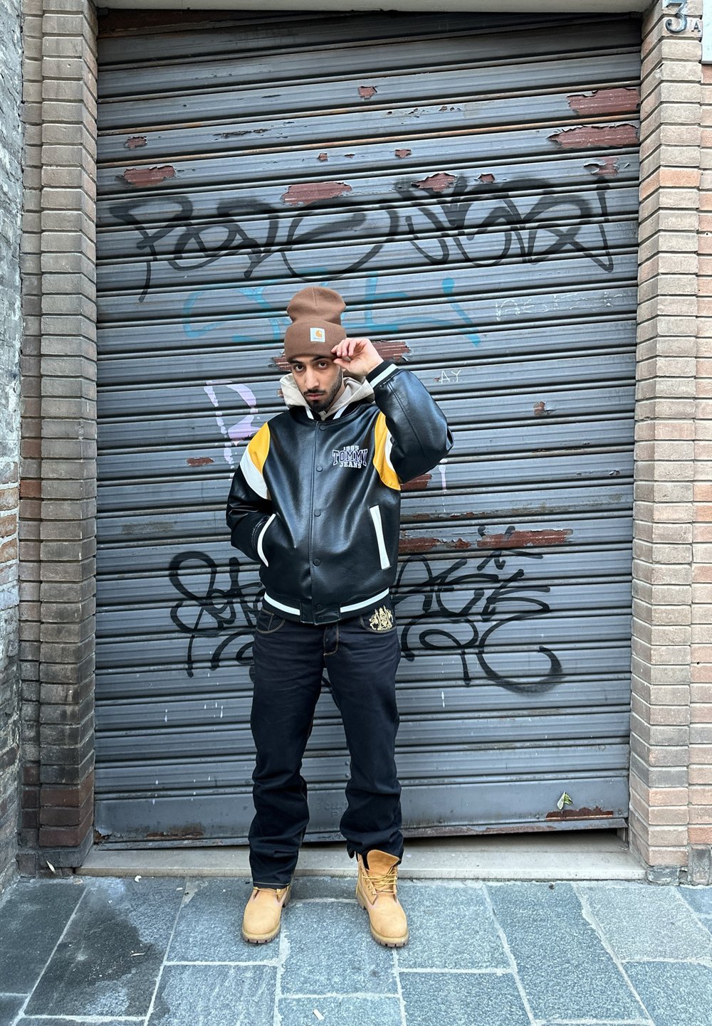 Μαύρο δερμάτινο μπουφάν bomber με κίτρινες και λευκές λεπτομέρειες, σκούρες τζιν παντελόνες και καφέ μπότες. Το φόντο έχει μια γκρι μεταλλική περσίδα με γκράφιτι.