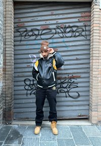 Μαύρο δερμάτινο μπουφάν bomber με κίτρινες και λευκές λεπτομέρειες, σκούρες τζιν παντελόνες και καφέ μπότες. Το φόντο έχει μια γκρι μεταλλική περσίδα με γκράφιτι.