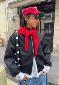 Femme portant une casquette rouge et une écharpe, des lunettes de soleil écaille de tortue, une veste noire zippée avec une chemise blanche, un jean bleu, et un sac noir à pois blancs.