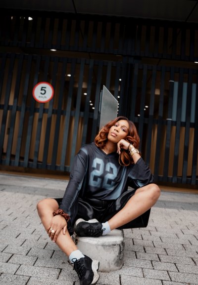 Jersey de gran tamaño en negro y gris con la impresión "22", combinado con pantalones cortos de cuero y zapatillas negras, sentado sobre un bloque de concreto.