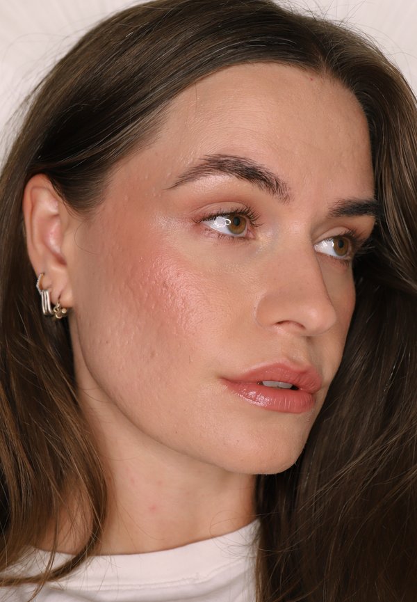 Gros plan d'une femme aux cheveux bruns, à la peau claire et au maquillage naturel, portant de petites boucles d'oreilles en or, regardant sur le côté sur un fond neutre.