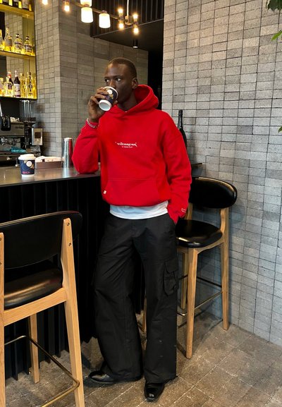 Sudadera roja con texto blanco, pantalones cargo negros y zapatos negros. De pie en una barra con una pared texturizada gris y un taburete de bar de madera.