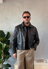 Czarna skórzana kurtka z zamkiem z przodu, kołnierzem i kieszeniami, w połączeniu z czarnym T-shirtem i kraciastymi spodniami. Wnętrze z roślinnością.