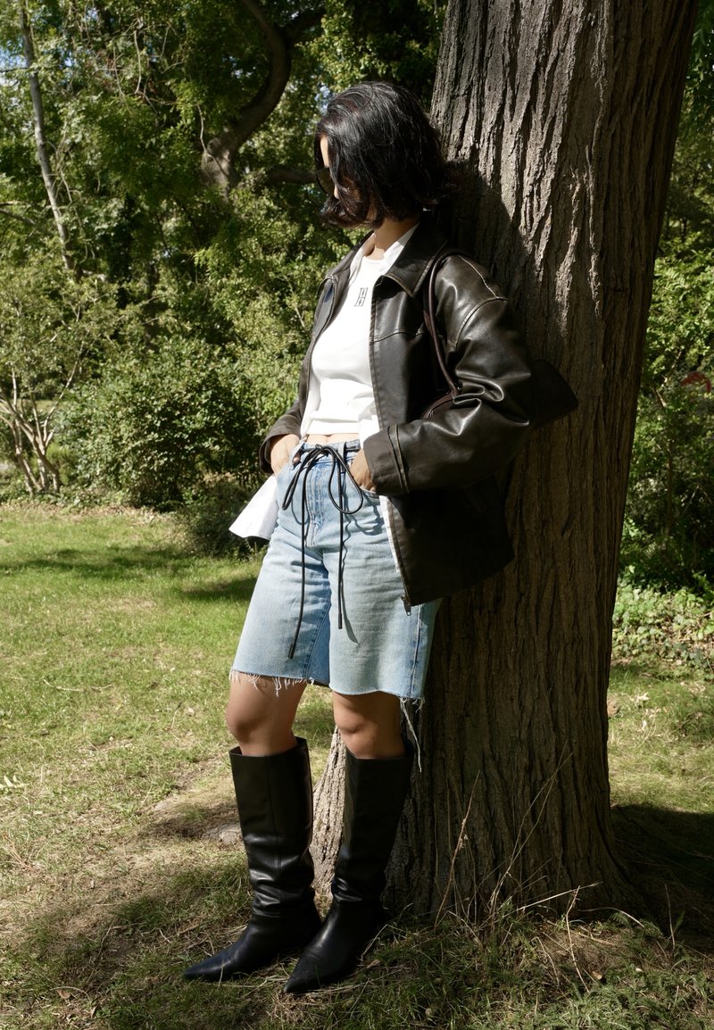 Giacca di pelle nera, top bianco corto, pantaloni denim blu con orlo sfrangiato e stivali neri fino al ginocchio. La modella è appoggiata a un albero.