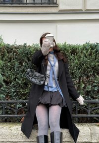 Črni plašč, siv pulover, modra ruta, črna torbica, puhaste rokavice. Drži skodelico kave, sedi pred zelenim grmičjem in kamnito ograjo.