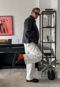 Czarna skórzana kurtka na czarnej koszuli, białe spodnie i czarne buty z akcentem w postaci pomarańczowych skarpet. Stojący obok regału i dzieła sztuki.