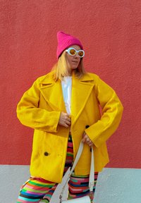 Gele wollen jas met oversized ontwerp, gecombineerd met gestreepte broek in meerdere kleuren. Accessoires zijn onder andere een roze gebreide muts en een witte handtas.