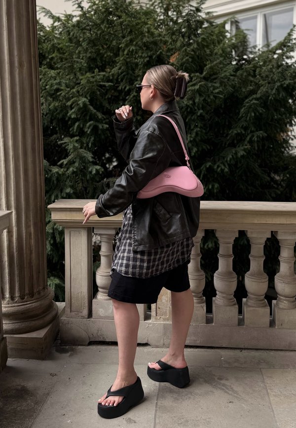 Personne debout sur une terrasse en pierre près d'une colonne et d'une rambarde, portant une veste en cuir noire, une chemise à carreaux, un short noir, des sandales à plateforme et portant un sac rose.
