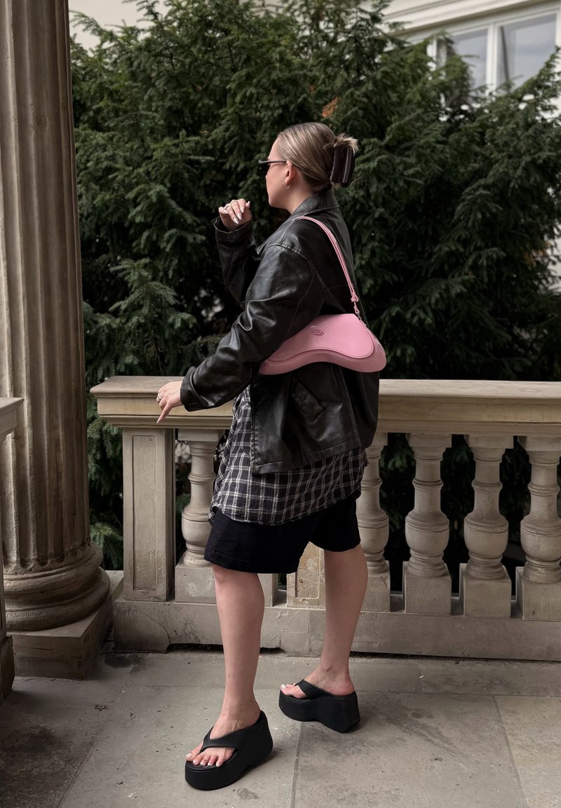 Personne debout sur une terrasse en pierre près d'une colonne et d'une rambarde, portant une veste en cuir noire, une chemise à carreaux, un short noir, des sandales à plateforme et portant un sac rose.