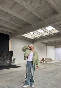 Zelená prošívaná bomber bunda, bílé tričko, roztrhané volné džíny a černobílé slip-on boty s kostkovaným vzorem. Průmyslové minimalistické prostředí.