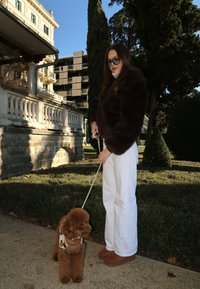 Femme portant une veste en fourrure foncée, un pantalon blanc et des lunettes de soleil tenant une laisse avec un petit caniche brun dans un jardin près d'un grand bâtiment.