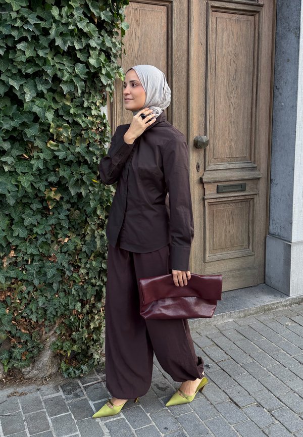 Femme portant un hijab beige et une tenue marron foncé se tient près d’un mur couvert de lierre et d’une porte en bois, tenant une grande pochette bordeaux et portant des talons jaunes.