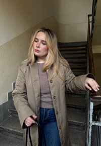 Lysebrun, lang frakke med knapper og glat tekstur, båret over en tætsiddende beige top og blå jeans, kombineret med en sort taske.