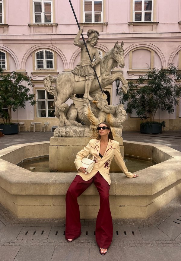 Mujer con gafas de sol, chaqueta beige y pantalones rojos sentada en el borde de una fuente de piedra histórica con una estatua de un guerrero a caballo.