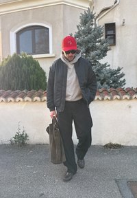 Veste bomber noire par-dessus un sweat à capuche gris, pantalon noir décontracté, chaussures marron, et tenant un sac marron. Arbuste vert et bâtiment en arrière-plan.