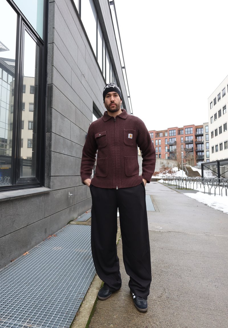 Mann iført en burgunderrød genser med glidelås og svarte bukser står på en fortau i byen nær moderne bygninger på en snøfylt dag.